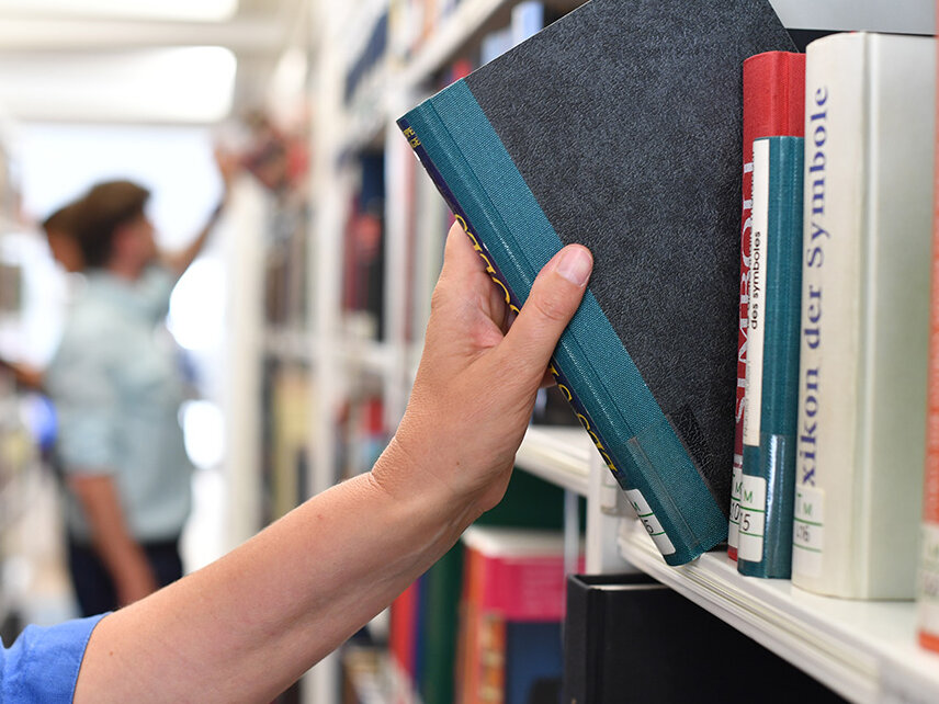 © Foto: Uli Deck Eine Hand zieht gerade ein Buch aus einem Bücherregal. Im Hintergrund ist eine Person zu sehen, die ebenfalls ein Buch aus dem Bücherregal entnimmt.