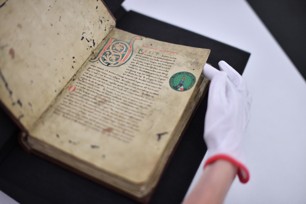 © Foto: Uli Deck Das Bild zeigt eine Mitarbeiterin der BLB, die mit Handschuhen die Nibelungenlied-Handschrift C (Cod. Hs. Don. 63) aus dem 2. Viertel des 13. Jahrhunderts aufschlägt.