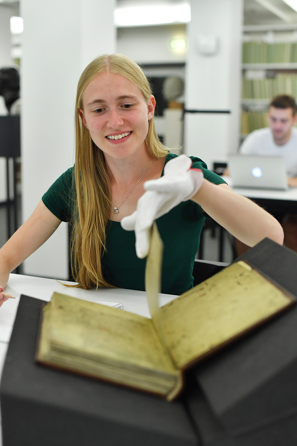 © Foto: Uli Deck Zu sehen ist ein aufgeschlagenes Buch im Lesesaal Sammlungen. Eine junge Frau blättert in diesem historischen Buch gerade eine Seite um. Sie lächelt und trägt weiße Handschuhe.